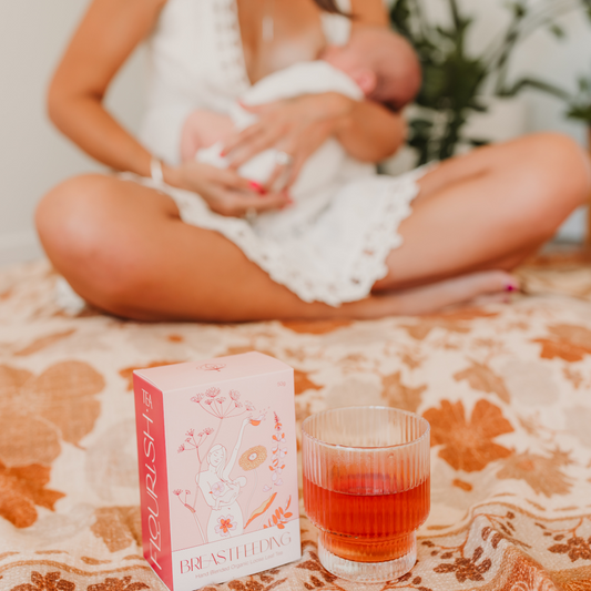 Woman breastfeeding a baby with a glass of amber liquid and a box labeled 'Breastfeeding' on a patterned surface.