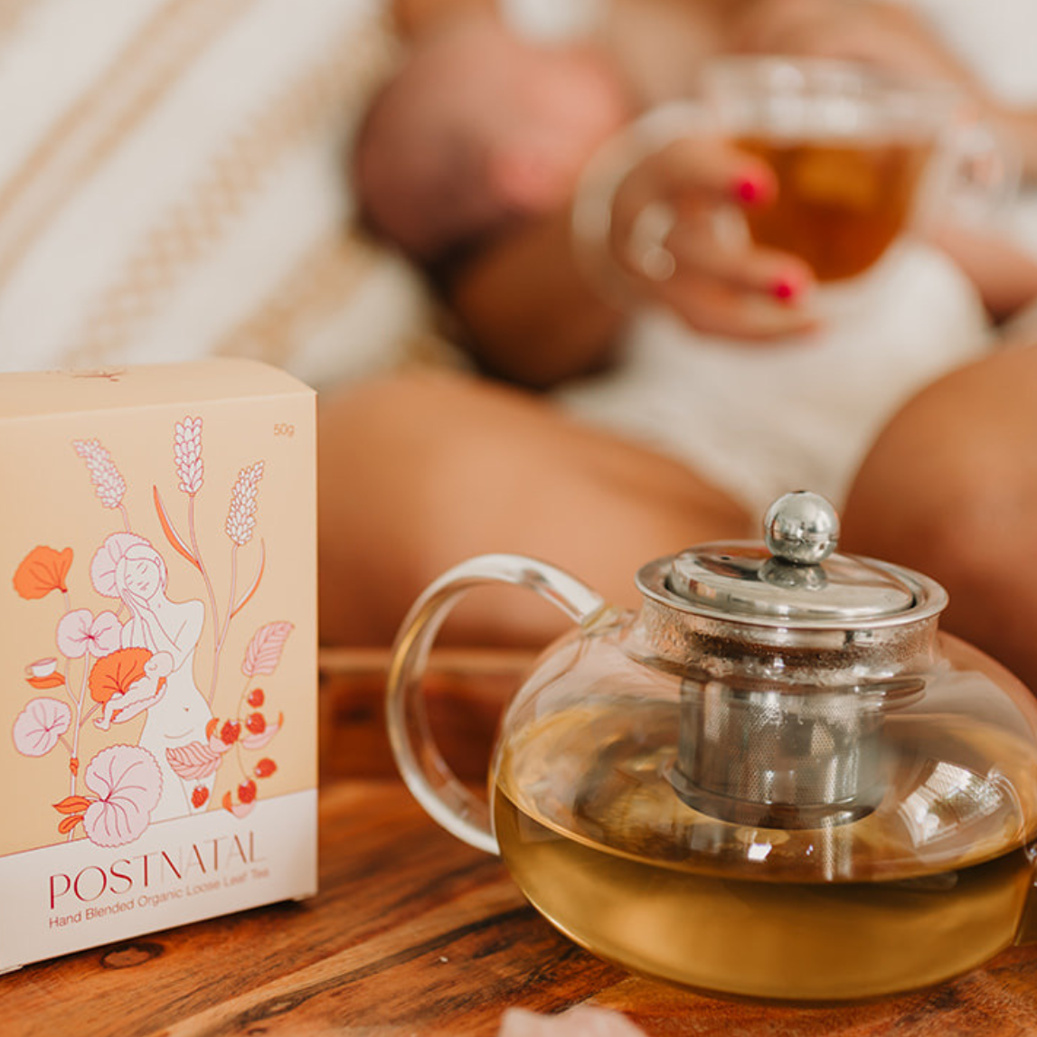 Glass teapot with tea on a wooden surface, with a blurred background of a person holding a cup.