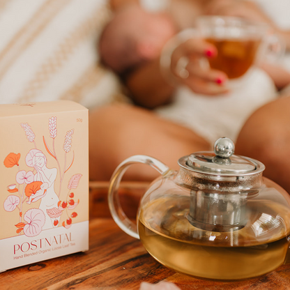 Glass teapot with tea on a wooden surface, with a blurred background of a person holding a cup.