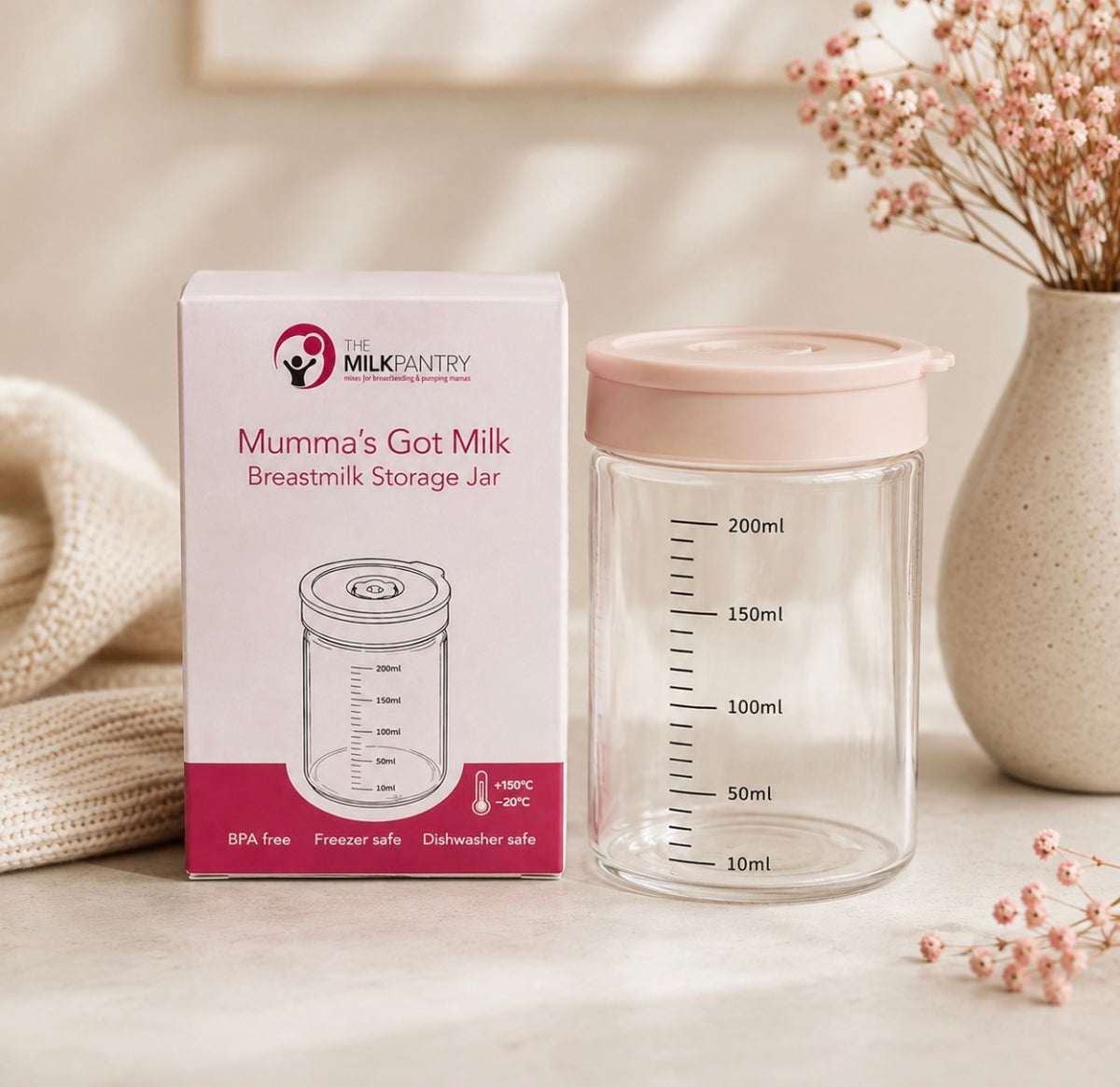 A clear breastmilk storage jar with a pale pink lid stands beside its branded retail box. The scene is styled on a light surface with soft beige props, including a textured blanket and a ceramic vase with dried flowers.