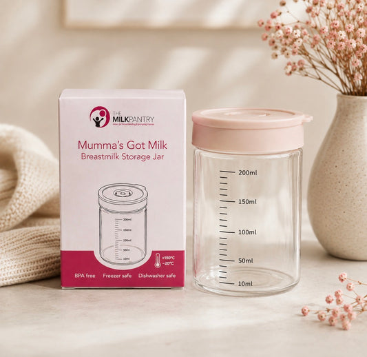 A clear breastmilk storage jar with a pale pink lid stands beside its branded retail box. The scene is styled on a light surface with soft beige props, including a textured blanket and a ceramic vase with dried flowers.