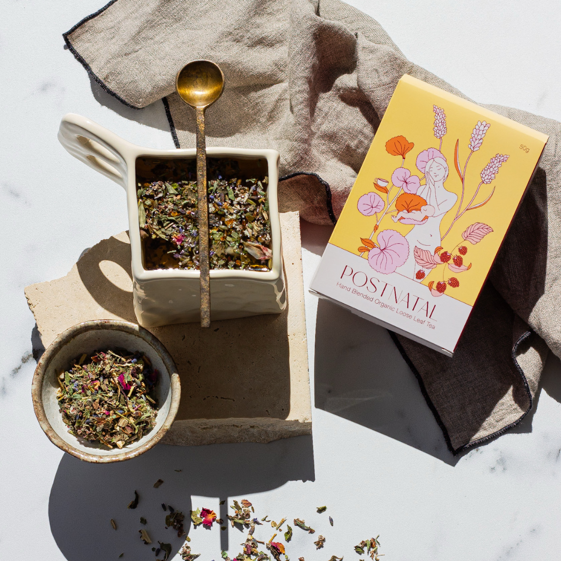 Tea-making setup with a teapot, bowls of tea leaves, and a box labeled 'Postimal' on a marble surface.