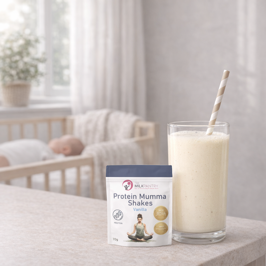 Glass of protein shake with a container labeled 'Protein Mumma Shakes' on a surface, with a blurred background of a room.