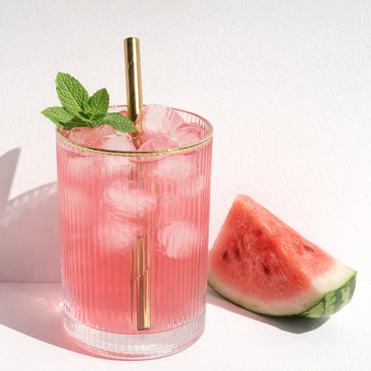 Pink cocktail with ice and mint in a glass with a gold straw, next to a slice of watermelon on a white background.