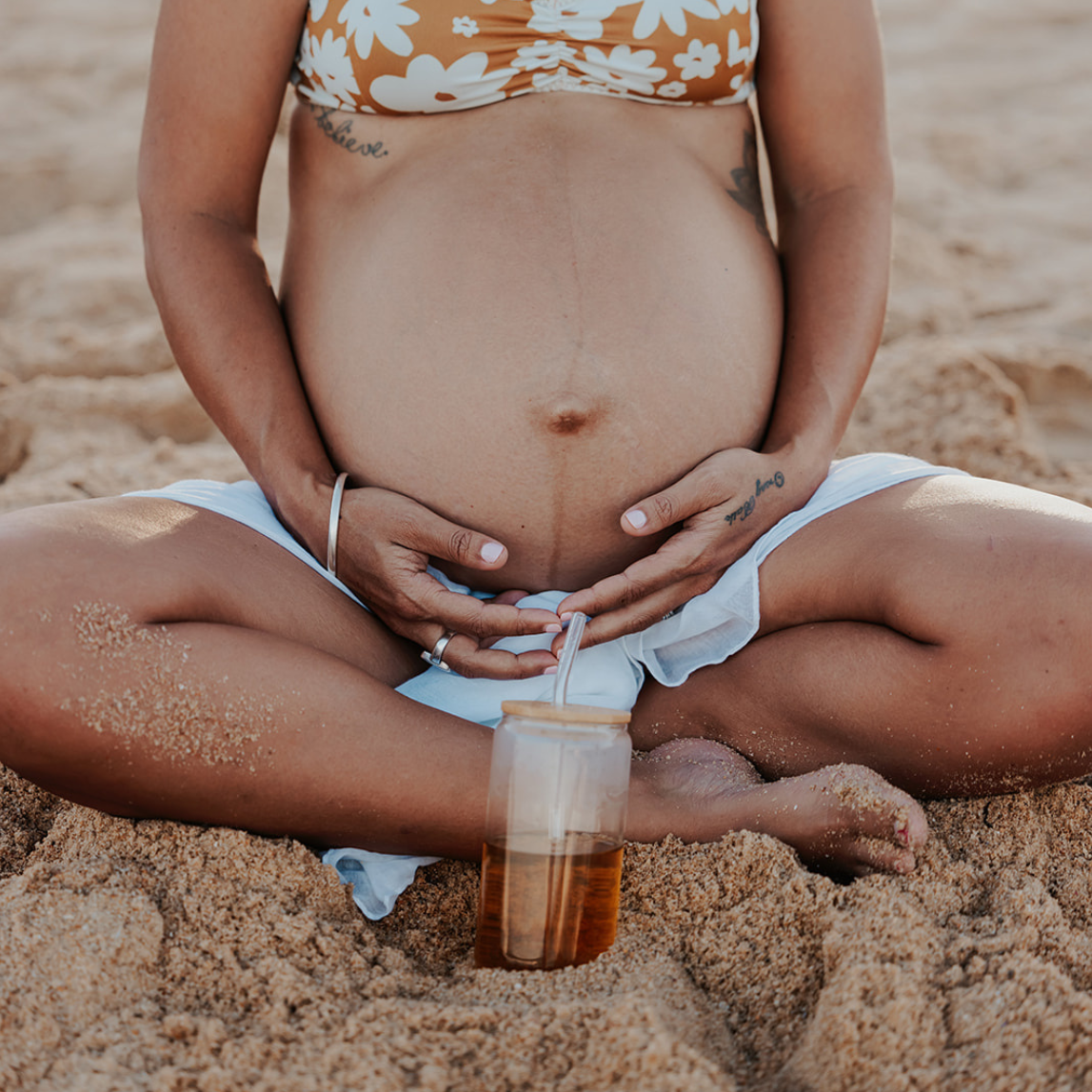 Pregnant woman sitting on the beach with a drink, wearing a floral bikini.