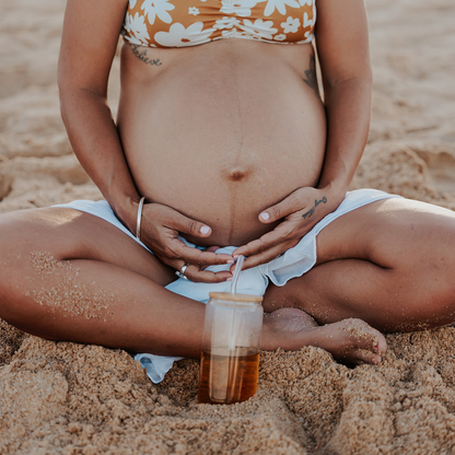 Pregnant woman sitting on the beach with a drink, wearing a floral bikini.